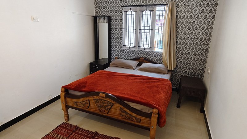 Bedroom with wooden double bed, red blanket, pillows, side table and window