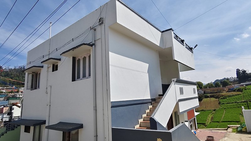 White cottage building with staircase beside green tea plantation and hillside houses under blue sky